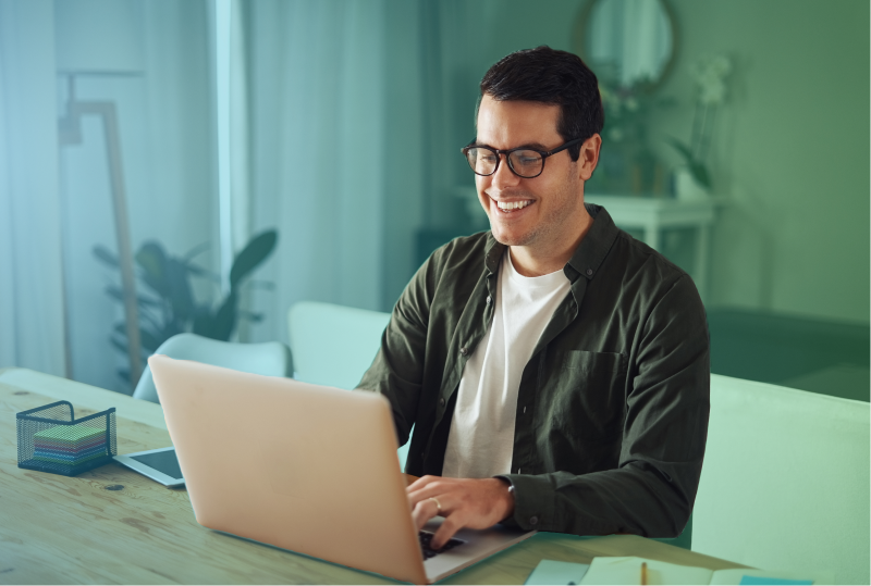 An employee is sitting at their desk in their home office, looking at their laptop and smiling.