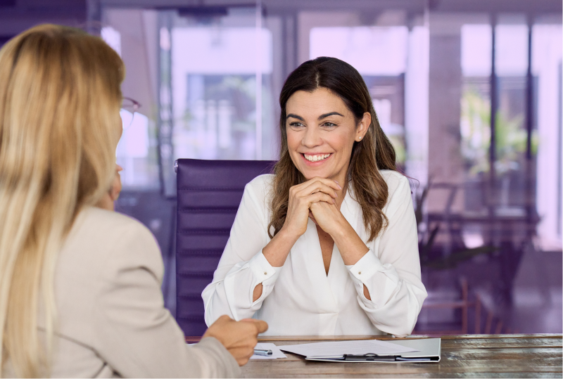 Two employees are sitting across a desk from each other, smiling and talking about their hiring strategy.