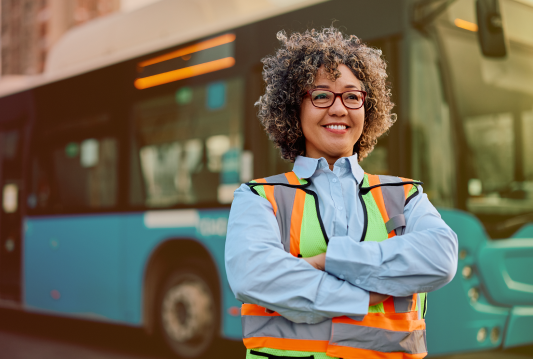 An employee in a safety vest is smiling and standing in front of a city bus/
